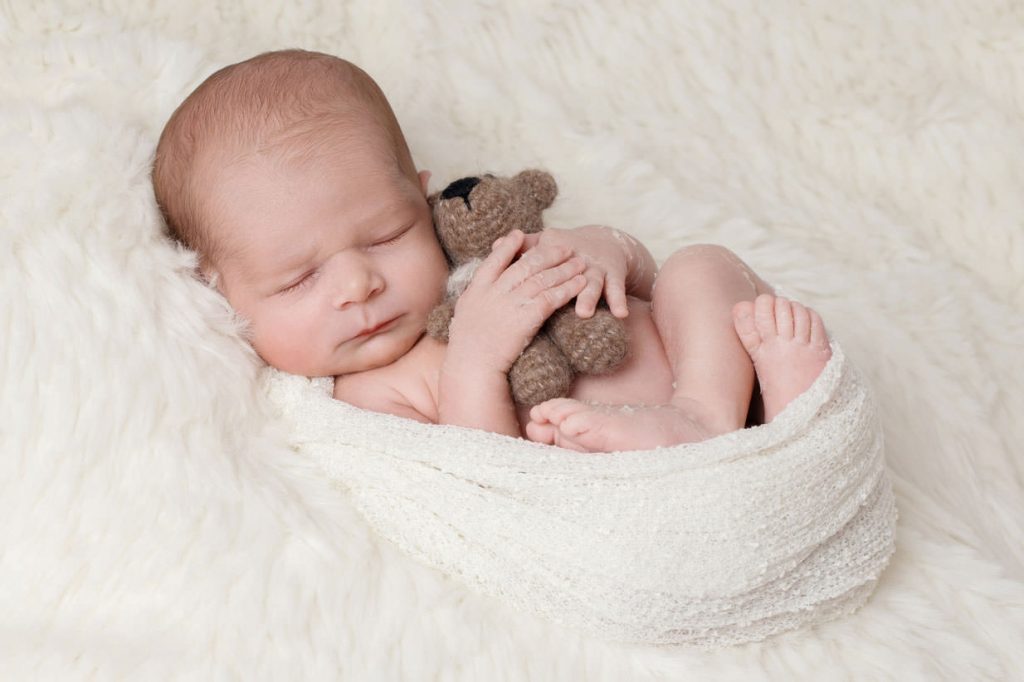 schlafendes, mit einem tuch eng eingewickeltes baby. auf seiner brust hält es einen kleinen gestrickten teddybär