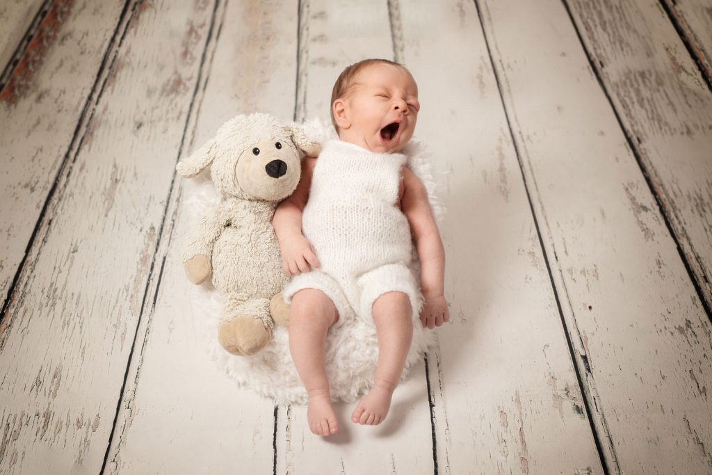 gähnend sitzt das baby angelehnt in einem sessel. neben ihm sein kuschelschaf